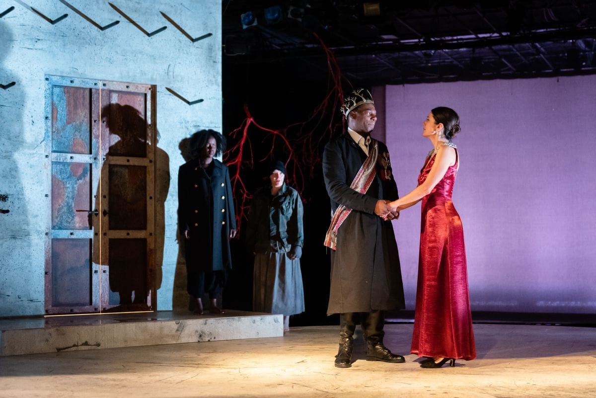 Two people watch on as Macbeth holds hands with lady Macbeth outside a castle.