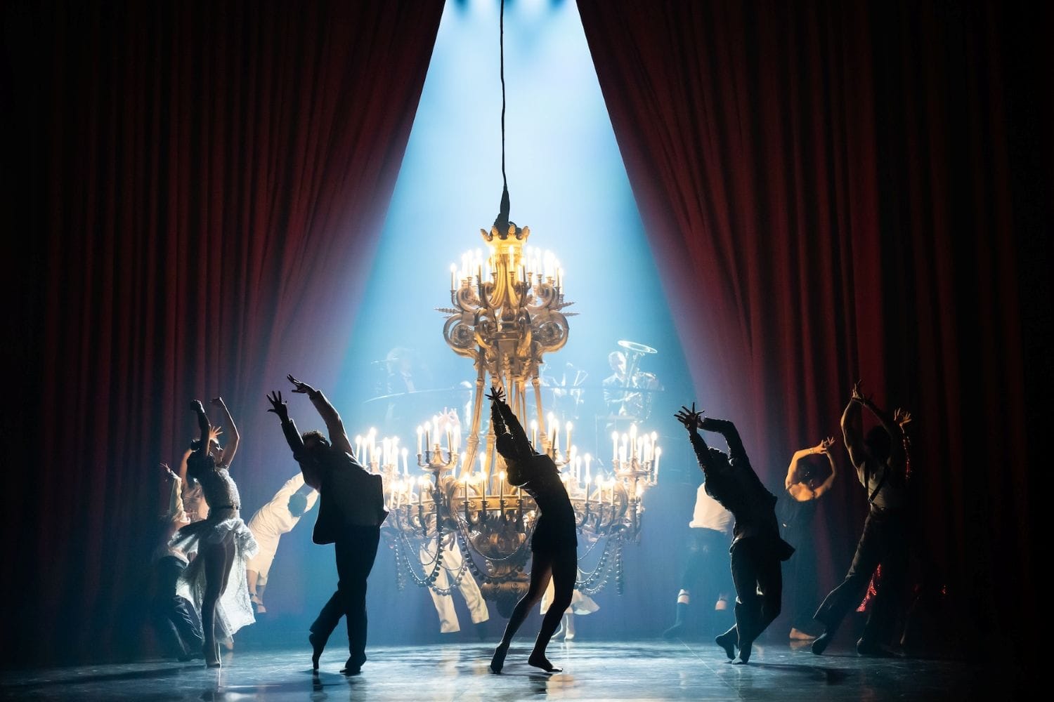 Dancers, some in shadow, dance onstage around a large chandelier that is close to the floor.
