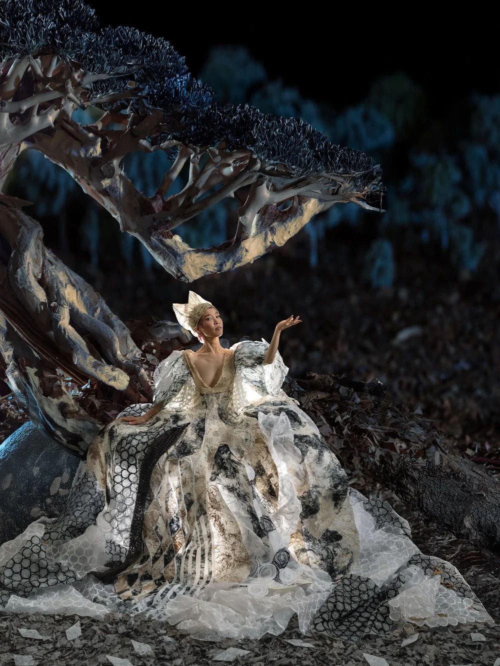 A woman in an dress made of multiple fabrics, aesthetics, and textiles stands regally in front of a large tree.