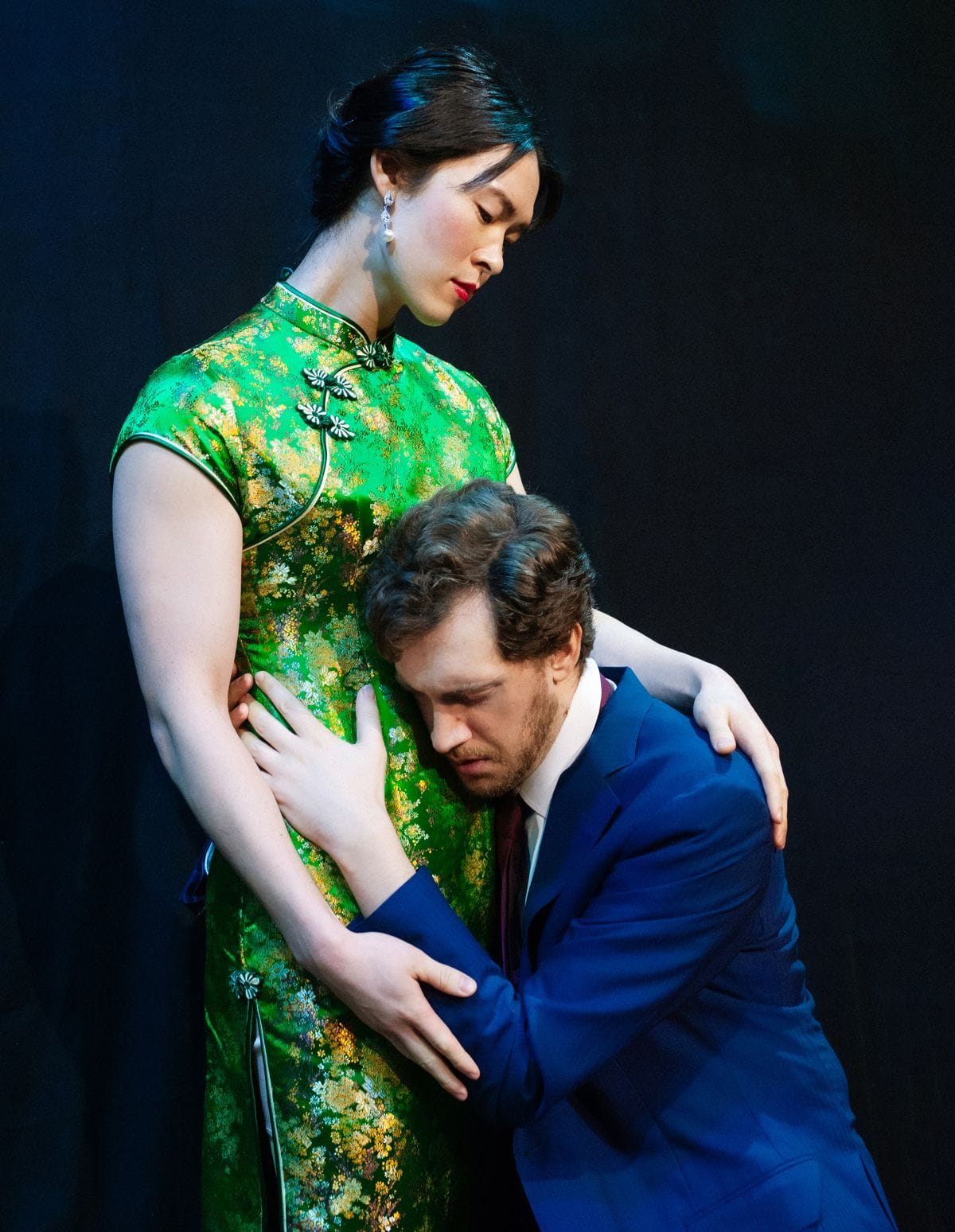 A women stands in a green Qipao gown. A man is on his knees holding her around her midsection as he pleads with her.
