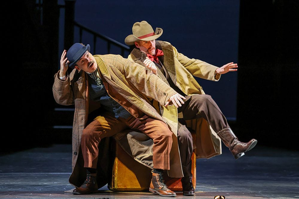Two men sit on a suitcase and they are falling off and over one another looking surprised.