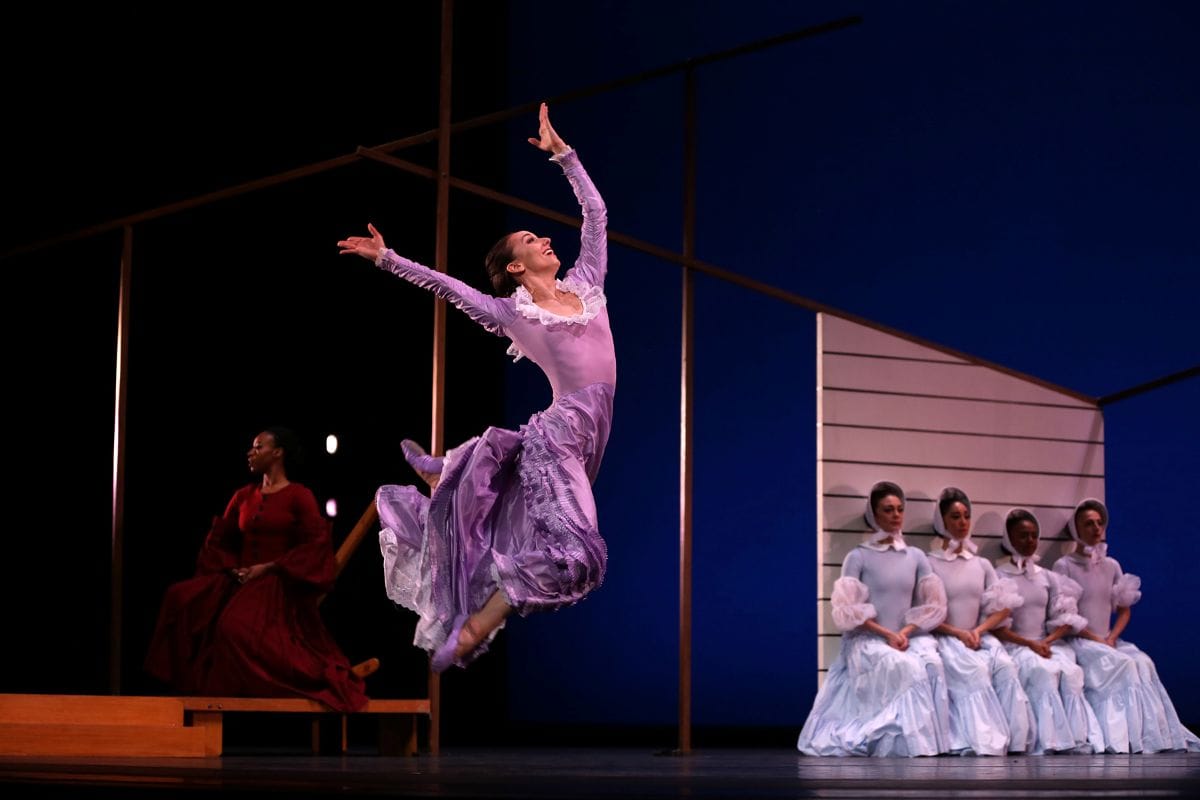 In the forefront a woman in a purple dress leaps into the air enthusiastically. Behind her, four women sit primly looking away.