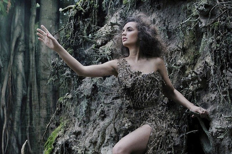 A woman stands in front of a tree, wearing a dress that matches the bark. She looks off in the distance.
