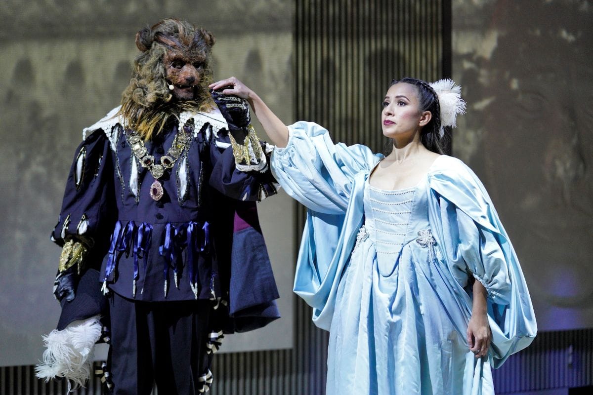 A man dressed as the beast with a mask and regal clothing holds the hand of a woman in a blue ball gown.