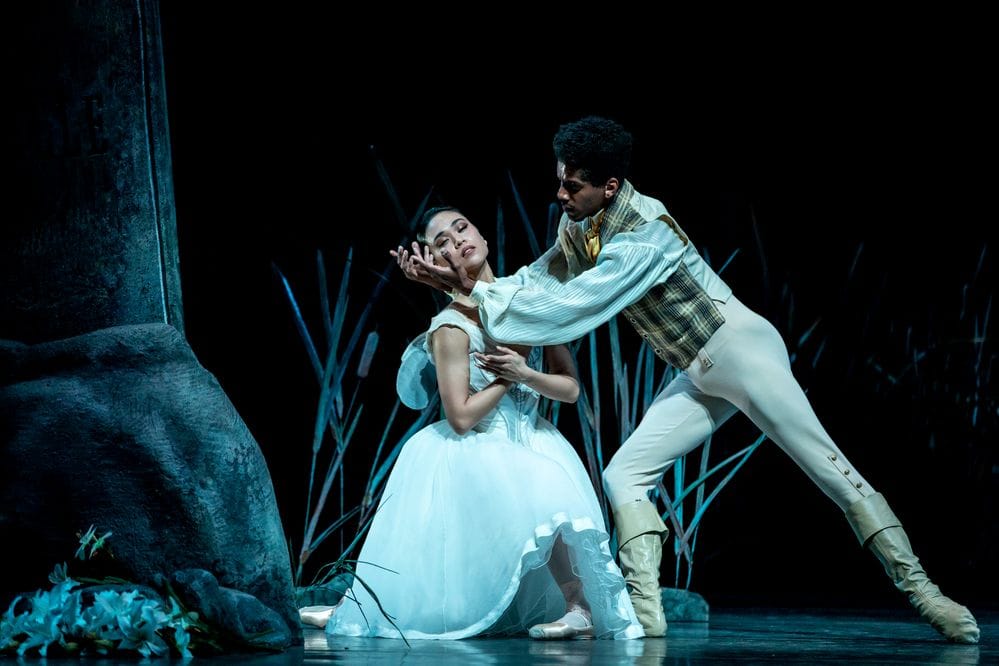 A ballerina knees with her eyes close as a man holds her face. They are cast in blue light.