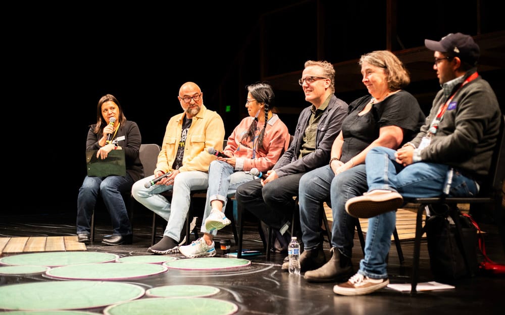 On stage a group of six people sit on chairs with microphones and speak in a panel setting.