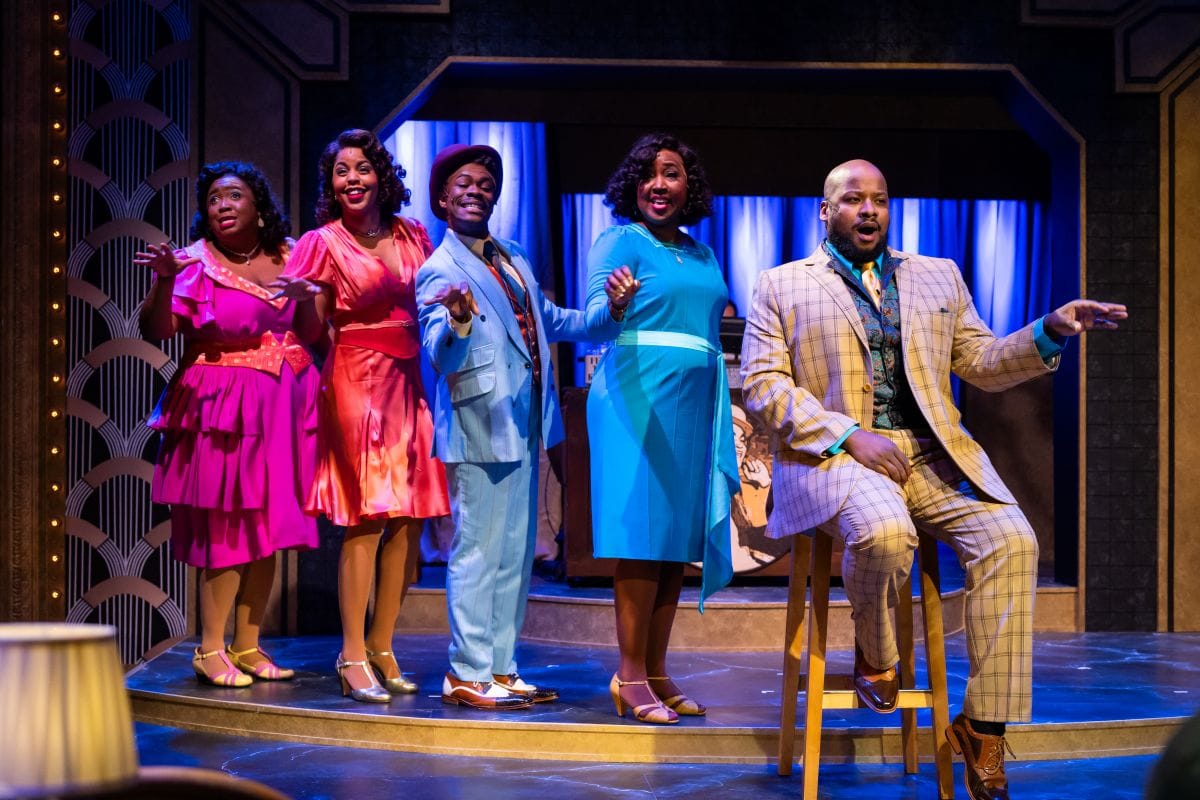 A group of men and women stand on a stage singing to the audience.