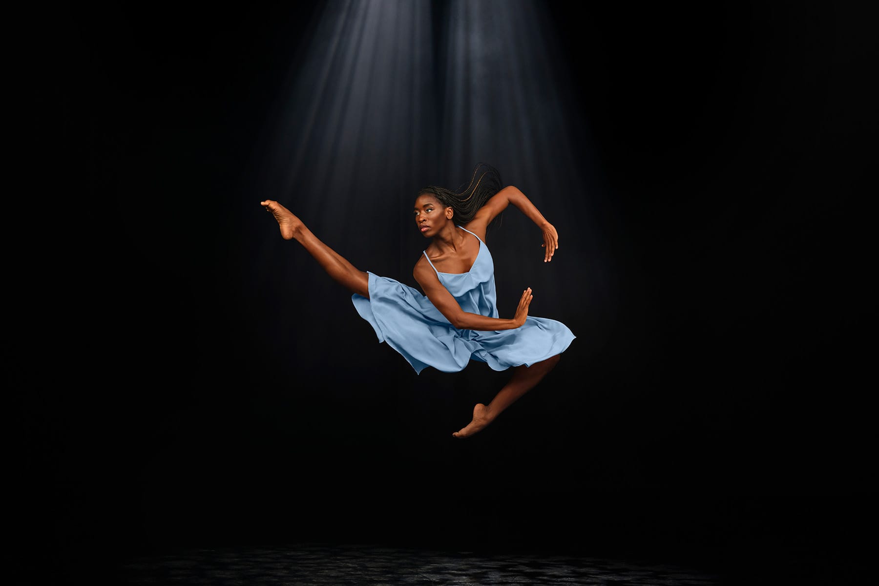 A ballet dancer in a blue dress is in a spotlight as she leaps with one leg extended against a black background.