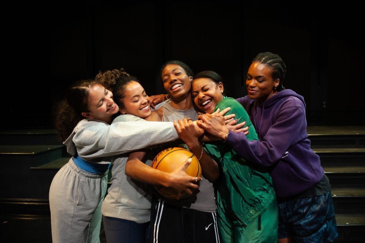 Teenage girls as members of a basketball team embrace together while the one in the middle holds a ball.
