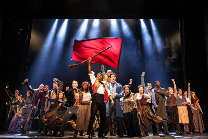 A large group of people stand with their arms in the air. A man in front holds a flag and a gun.