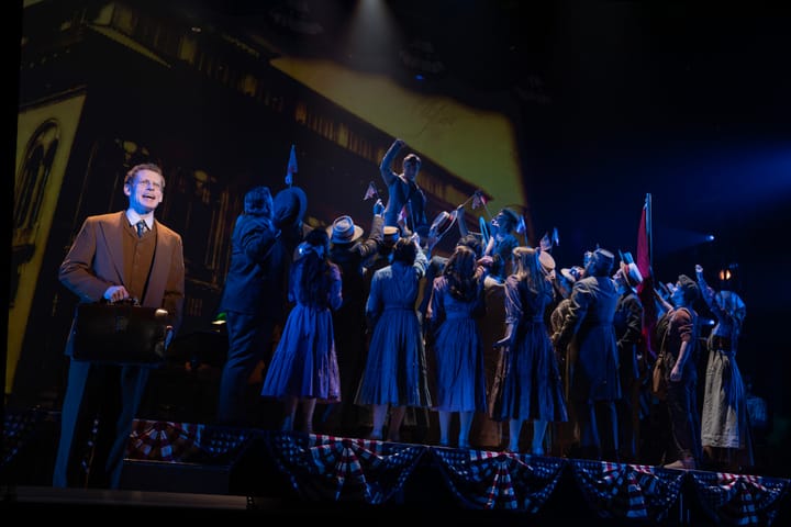 A man in the forefront sings with a spotlight while a large group of people in the background listen to a speaker.
