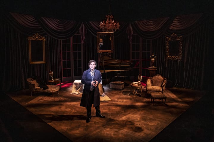 A man wearing 19th century clothes stands on a stage decorated as a living room from the same time period.