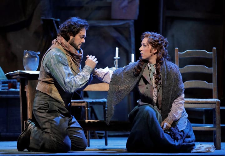 A man and woman kneel in front of a table. The man holds the woman's hand.