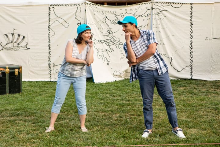 A man and woman stand looking at each other with their hands on their chins. They perform outside in front of a canvas backdrop.