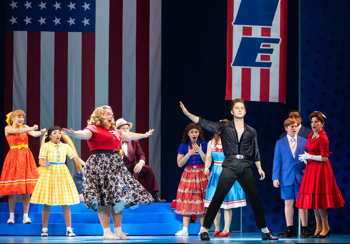 A man in black stands surrounded by fawning teen girls, all dressed in 50s outfits.