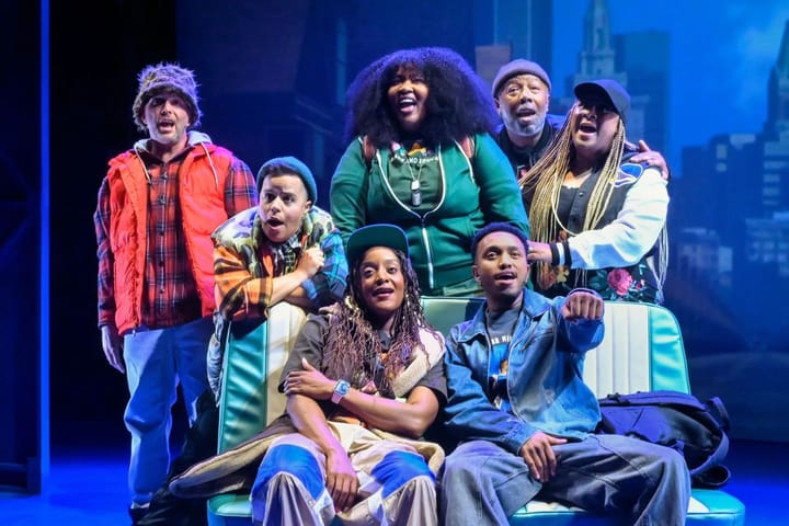 Two young people sit on a seat from a car while five others stand behind them. They all sing.
