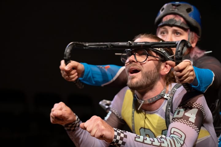 A woman with a helmet holds a bicycle bar against a mans forehead, looking like he's the bike.