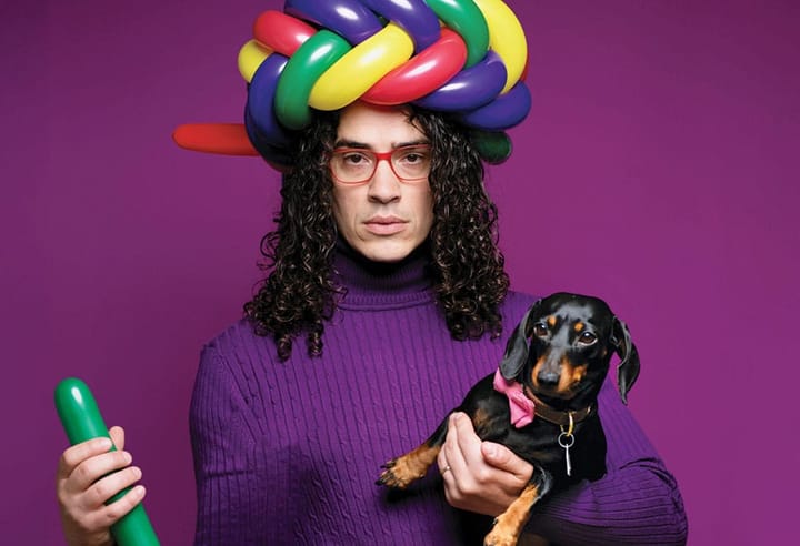 A man wears a purple sweater with a balloon crown on his head and holding a dog.
