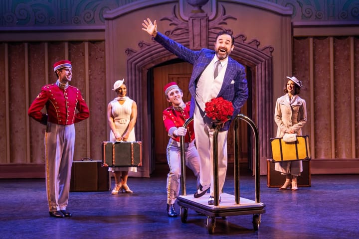 A set shows a hotel, with two bell hops in uniform. One pushes a man holding roses on a luggage cart.
