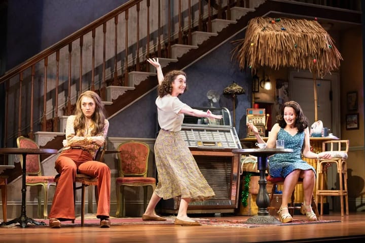 A set shows a living room in the 1970s. Two women sit, one mad, the other laughing, and a third dances.