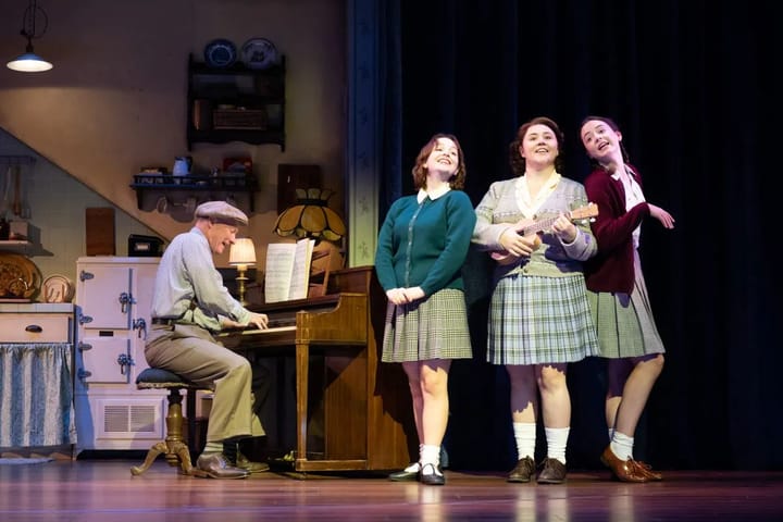Three women stand close together singing, the one in the middle plays the guitar. An older man plays the piano.