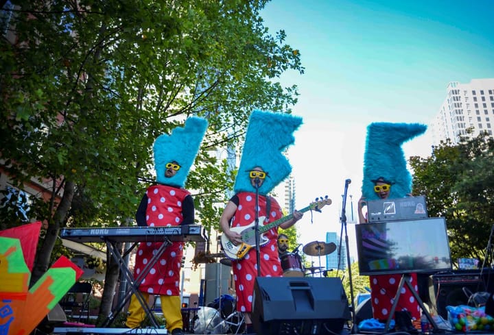 Three people wearing fuzzy blue headresses and polka dot dresses play instruments in a concert outdoors.
