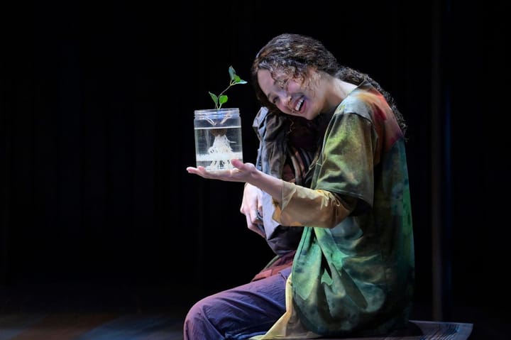 A woman kneels on the ground smiling. She is looking at a plant in a glass that is growing.