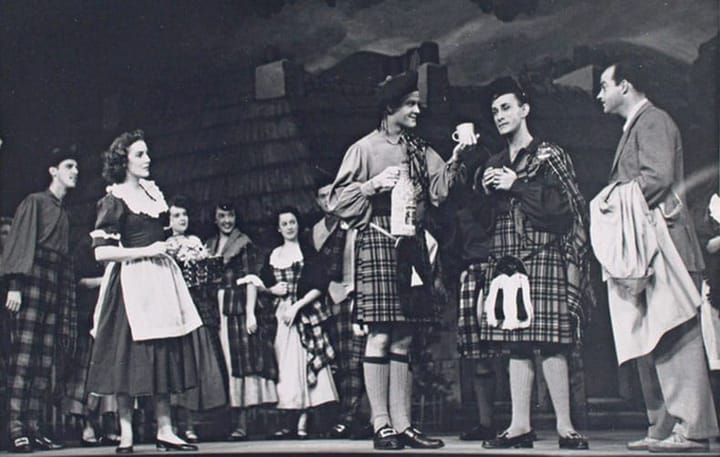 A black and white photo from the 1947 stage production of Brigadoon. Men wear kilts and women wear dresses with aprons.