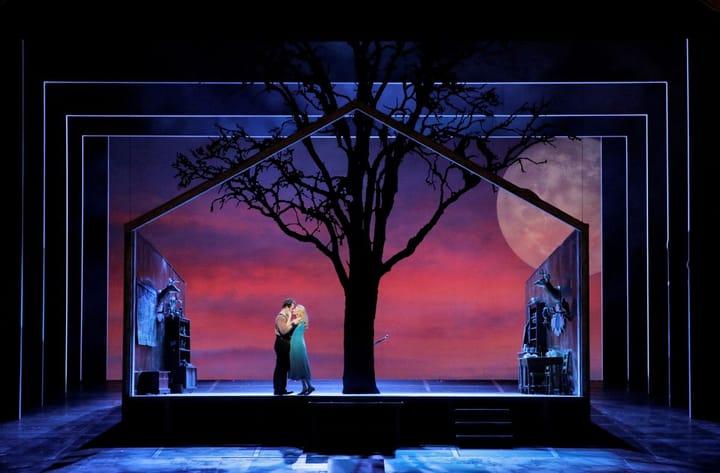 A large tree is in the center of the stage with a moon in the background. Two lovers embrace under the tree.