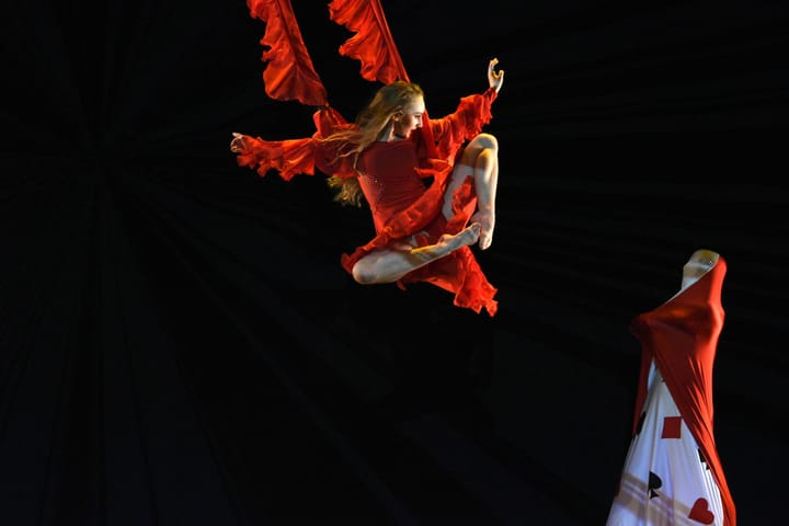 Against a completely black background, a dancer is doing an aerial stunt, flying at a woman dressed in a red and white outfit.