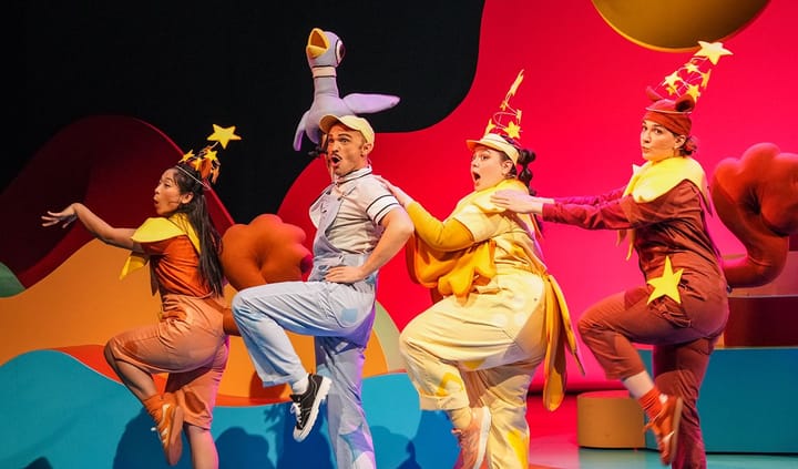 Four actors wearing colorful outfits are on a stage with stars on their hats and are jumping together.