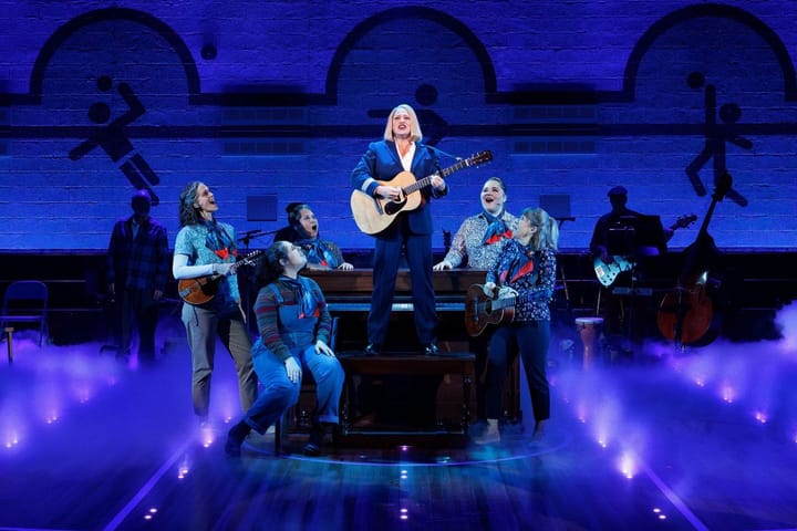 A woman playing the acoustic guitar stands on a piano bench singing with a group of women backing her up.