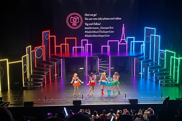 On stage a colorful light up set shows a skyline and on a screen are sing a long words. On stage are the sailor guardia