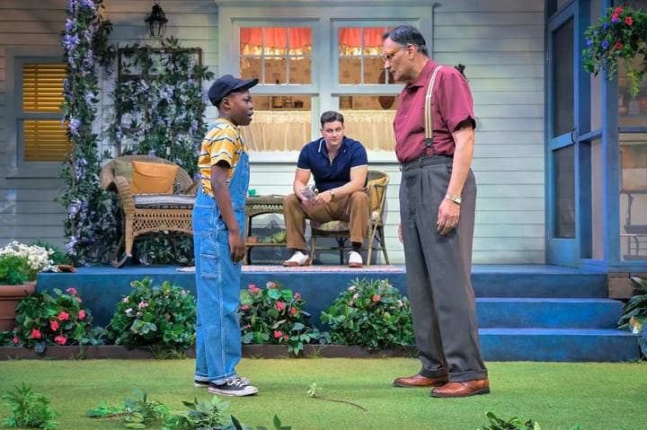 The set shows the backyard of a home. A man sits on the porch looking at a older man and young boy who are standing talking.