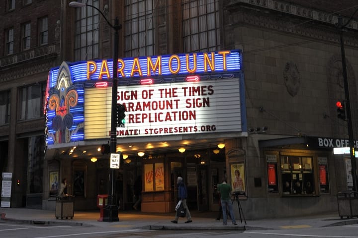 Exterior of a theatre, which is a large brick building with a marquee sign lit up. People walk outside on the sidewalk.