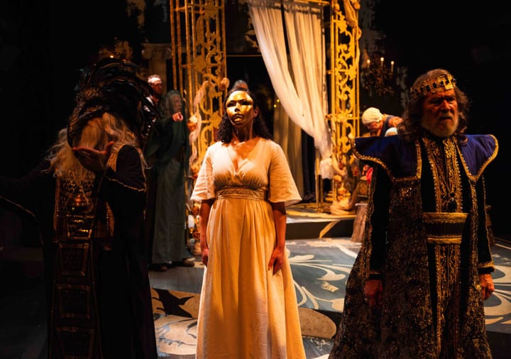 A woman in a white dress stands in the center wearing a gold mask over her face. A King stands to her right in royal robes.