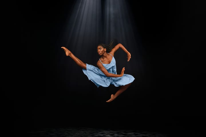 A ballerina wearing a blue dress leaps in the air with one leg extended, high off the ground against a black backdrop.
